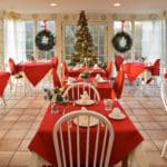 Spacious room surrounded by windows with several white dining tables draped in red cloths set for a meal at Christmas