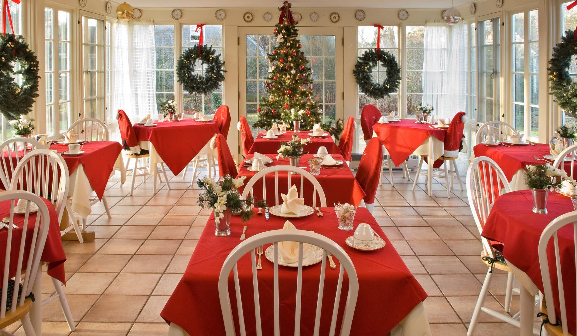 Spacious room surrounded by windows with several white dining tables draped in red cloths set for a meal at Christmas