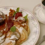 White plate trimmed in pale flowers topped with apple pancakes, powdered sugar, crispy bacon, strawberries and syrup
