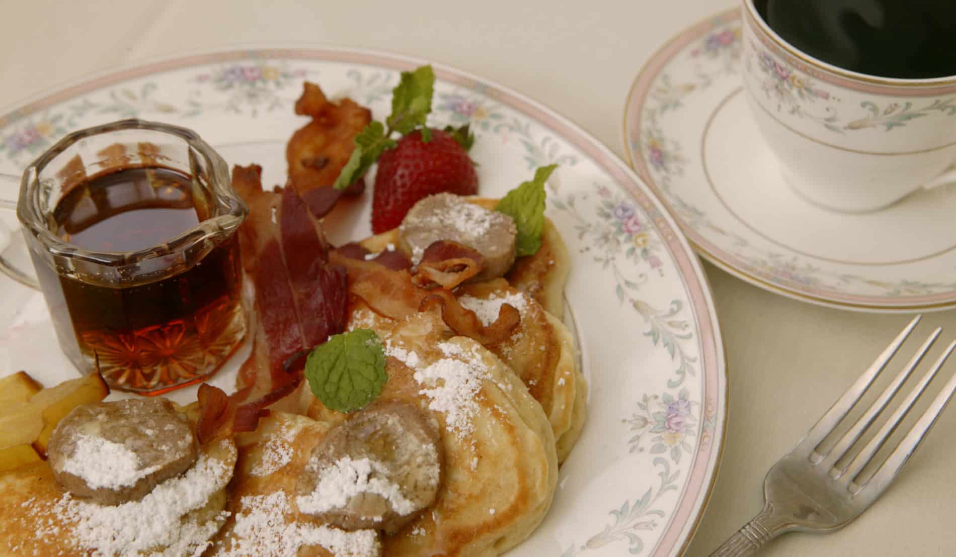 White plate trimmed in pale flowers topped with apple pancakes, powdered sugar, crispy bacon, strawberries and syrup