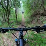 From the view of a mountain biker, bike handlebars, and another biker ahead on the wooded path.