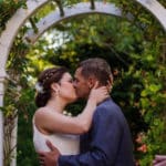 Bride and groom kissing under a white arched trellis with flowering vine