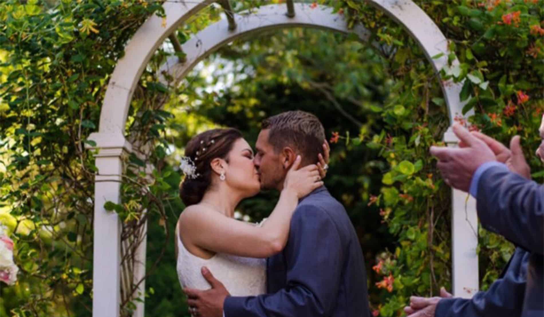 Bride and groom kissing under a white arched trellis with flowering vine