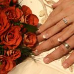 Groom and bride's hands with wedding rings alongside bouquet of red roses