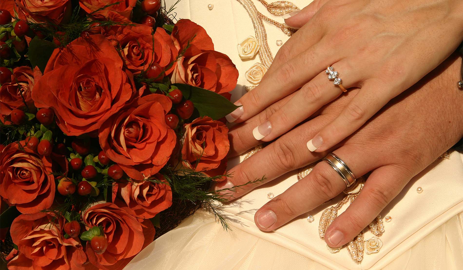 Groom and bride's hands with wedding rings alongside bouquet of red roses