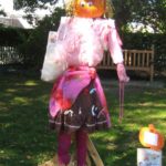 Female scarecrow in pink with a pumpkin head propped up in the grass