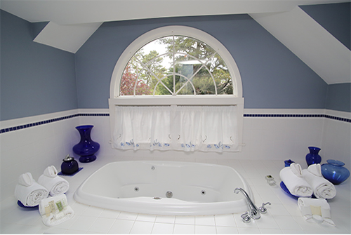 Beautiful pale blue and white bathroom with soaking tub, white tile and towels, blue vases and arched window