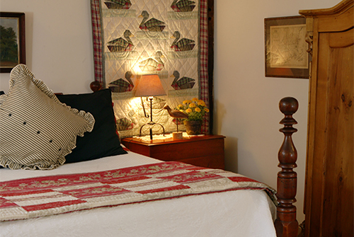 Whirlwind guest room showing queen bed with red quilted bedding, quilted wall hanging, and night side table with reading light