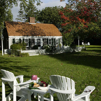 Two white Adirondack chairs and small table in the grass by a white sided cottage with white picket fence