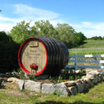 Large barrel that says Truro Vineyards on Cape Code surrounded by rocks, white fencing, greenery and blue skies